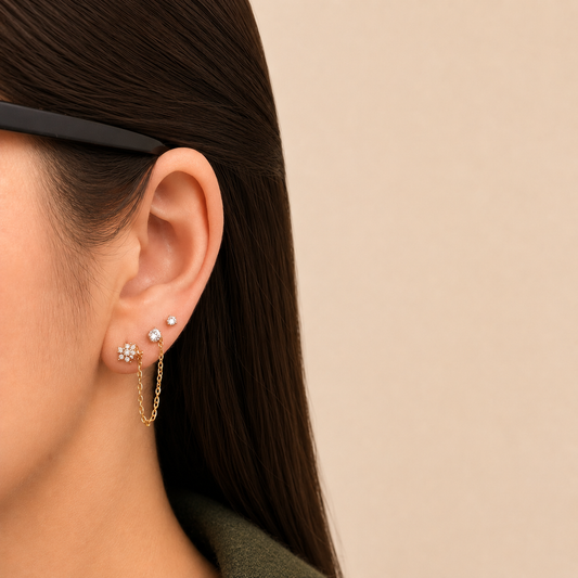 A close-up of a model's ear wearing a delicate gold huggie hoop earring with a small dangling chain and a flower-shaped stud, alongside small pearl studs, against a warm beige background.